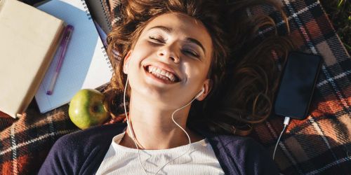 Studentessa, sdraiata su plaid, ascolta musica. Di fianco a lei un quaderno, un libro e una mela