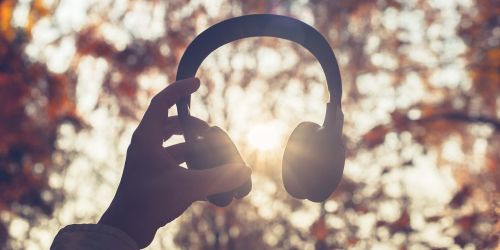 Mano che sorregge cuffie musicali, sullo sfondo fronde di alberi ad inizio autunno