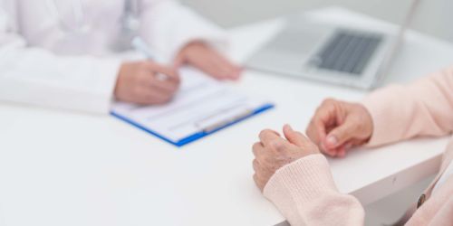 Dettaglio su scrivania a cui siedono paziente e medico che scrive su una cartelletta