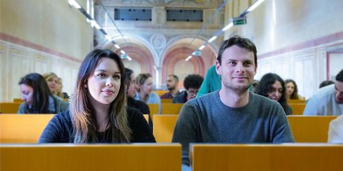 Una studentessa e uno studente in Aula. Sullo sfondo, sfuocati, altri studenti e studentesse