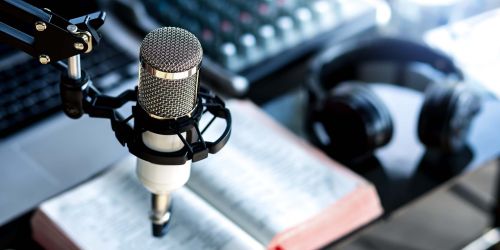 Microfono radiofonico in primo piano, sullo sfondo cuffie, un libro, un pc e un mixer