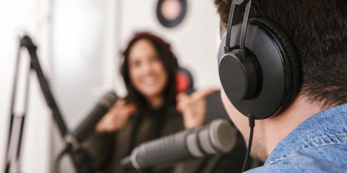 Registrazione di un podcast: uomo di spalle con cuffie al microfono, di fronte a lui giovane donna con con cuffie