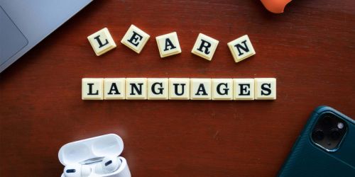 Tesserine formano la scritta "Learn languages" su una scrivania su cui sono poggiati anche un cellulare, delle cuffie audio e un pc