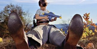 Scena dal film "La congiura degli innocenti" con bambino che regge pistola ad acqua e in primo piano i piedi di un uomo steso a terra