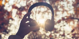 Mano che sorregge cuffie musicali, sullo sfondo fronde di alberi ad inizio autunno