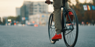 Dettaglio su gambe di uomo in sella a una bicicletta. Sullo sfondo un paesaggio cittadino.