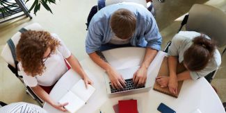 Due donne e un uomo seduti a un tavolo rotondo, visti dall'alto, mentre lavorano al pc con appunti