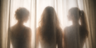 Sagome di tre donne di spalle dietro a una tenda bianca, affacciate a una finestra