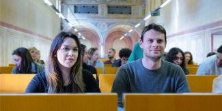 Una studentessa e uno studente in Aula. Sullo sfondo, sfuocati, altri studenti e studentesse