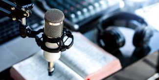 Microfono radiofonico in primo piano, sullo sfondo cuffie, un libro, un pc e un mixer